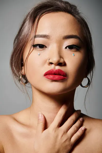 Retrato de mujer asiática con elegante maquillaje delicado tocando el cuello con la mano sobre fondo gris - foto de stock