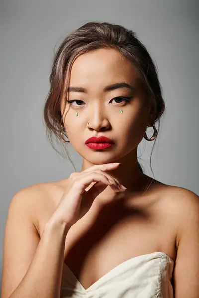 Портрет впевненої азіатки з елегантним макіяжем, який торкається підборіддя з рукою на сірому фоні — стокове фото