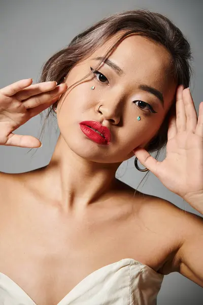 Портрет красивой азиатской женщины с красными губами, держащей руки вокруг лица на сером фоне — стоковое фото