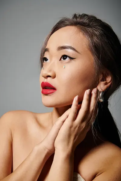 Retrato de elegante mulher asiática com lábios vermelhos mãos dobráveis e olhando para cima no fundo cinza — Fotografia de Stock