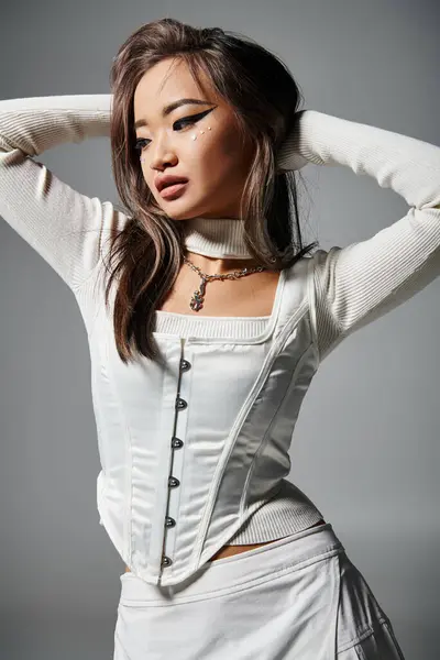 Bastante asiático mujer en elegante traje con atrevido maquillaje poner la mano detrás de la cabeza y mirando hacia abajo - foto de stock