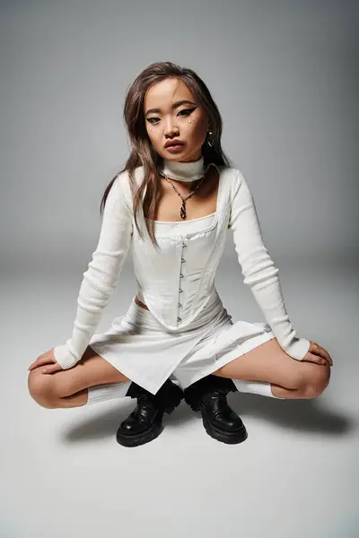 Привлекательная азиатская молодая женщина в стильном наряде присела на сером фоне — стоковое фото