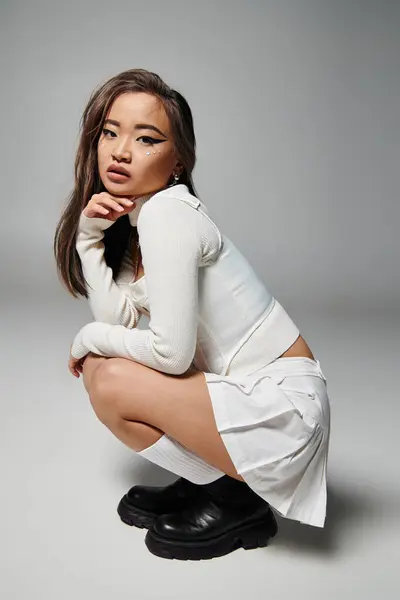 Азиатская женщина в белом стильном наряде приседает боком на сером фоне — стоковое фото