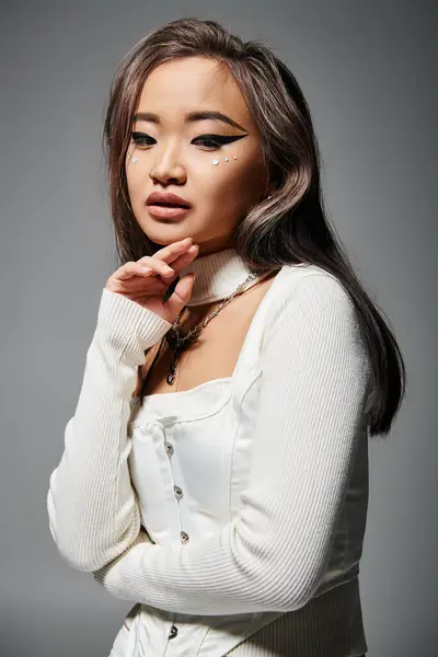 Hermosa asiático joven mujer con outrageous maquillaje delicado tocar a barbilla en gris fondo - foto de stock