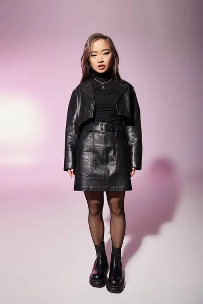 Atractiva mujer asiática en traje de cuero negro con maquillaje pesado de pie sobre fondo lila - foto de stock