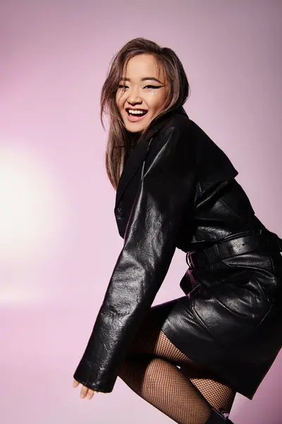 Sonriente asiático joven mujer en negro traje de cuero con pesado maquillaje posando sobre lila fondo - foto de stock