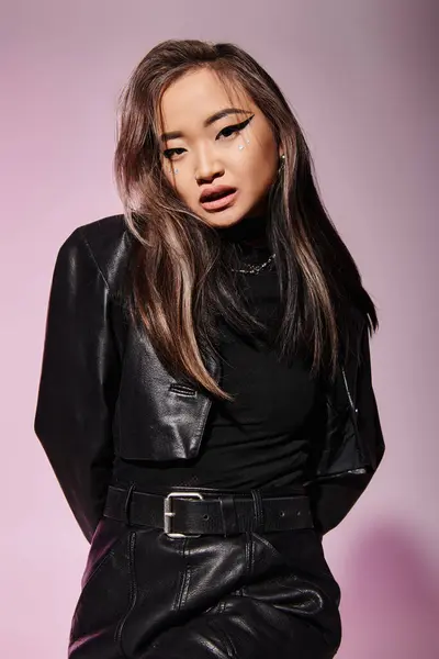 Hübsche asiatische Frau im schwarzen Leder-Outfit mit schwerem Make-up-Look vor der Kamera auf fliederfarbenem Hintergrund — Stockfoto