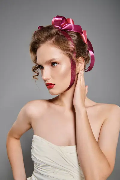 Молода жінка виявляє класичну красу в білій сукні з рожевим бантом на голові, елегантно позуючи в студії на сірому фоні. — стокове фото