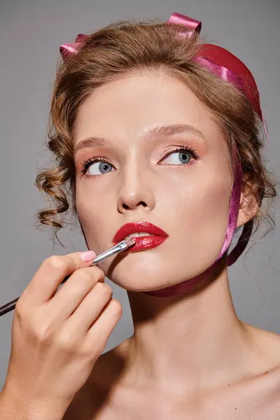 Uma jovem mulher com um laço rosa no cabelo está aplicando cuidadosamente batom em seus lábios em uma pose de beleza clássica em um fundo cinza.. — Fotografia de Stock