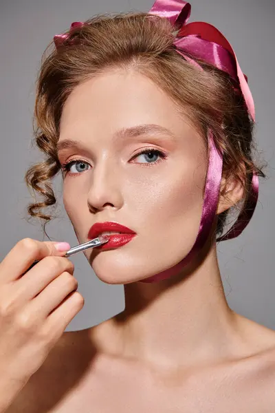 A young woman with a pink bow in her hair is applying lipstick to her lips, exuding classic beauty and elegance in a studio setting on a grey background. — Stock Photo