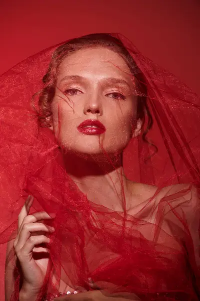 Молодая женщина излучает классическую красоту, позируя в студии в потрясающем красном платье с вуалем, каскадом над головой. — стоковое фото