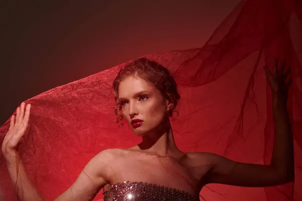 Una mujer joven exuda belleza clásica en un vestido sin tirantes, sosteniendo un paño rojo vibrante mientras que golpea una pose en un ajuste del estudio sobre un fondo negro. - foto de stock
