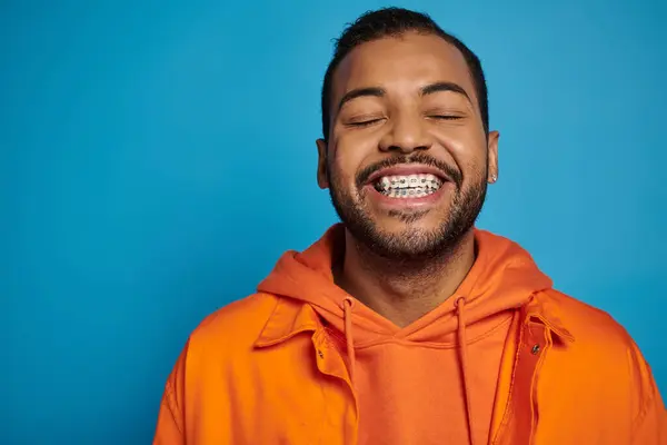 Encantador hombre afroamericano en sus 20 años sonriendo ampliamente con los ojos cerrados contra el fondo azul - foto de stock
