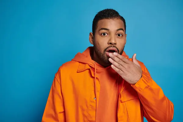 Hombre afroamericano sorprendido en traje naranja que cubre la boca con la mano sobre fondo azul - foto de stock