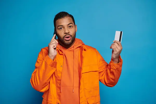 Surprised african american young guy calling in smartphone with credit card on blue background — Stock Photo