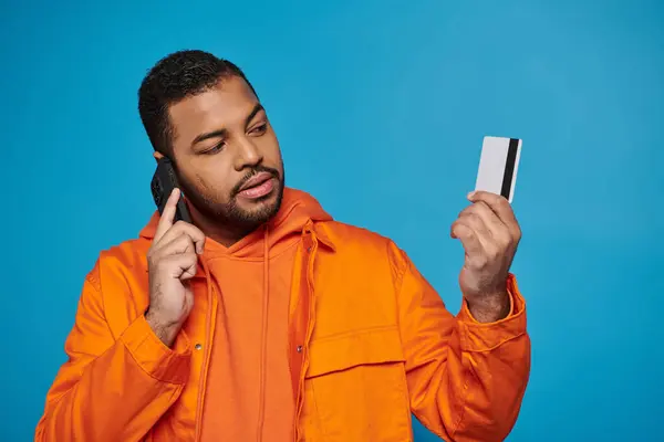 Очаровательный африканский американец в оранжевом костюме звонит в смартфон и ищет кредитную карту — стоковое фото