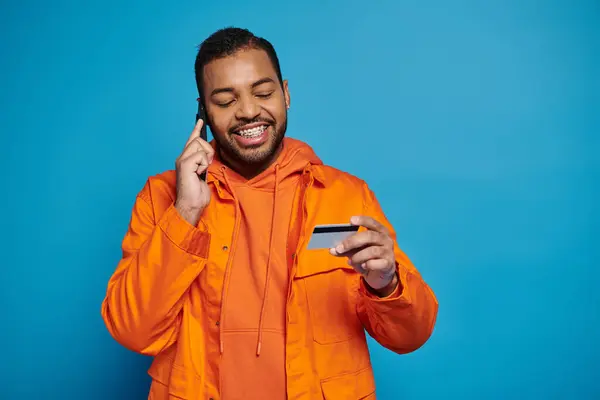 Feliz sonriente afroamericano hombre llamando en el teléfono inteligente y mirando la tarjeta de crédito en azul - foto de stock