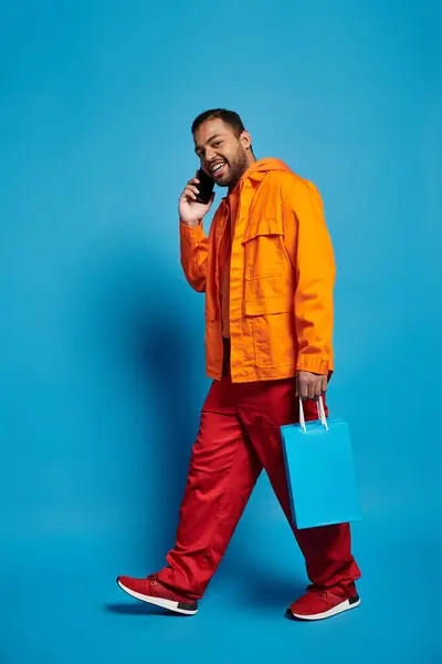 Atractivo afroamericano hombre llamando en smartphone y caminando con bolsas de compras - foto de stock