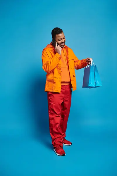 Charmanter afrikanisch-amerikanischer Mann ruft Smartphone und blickt auf Einkaufstüten auf blauem Hintergrund — Stockfoto