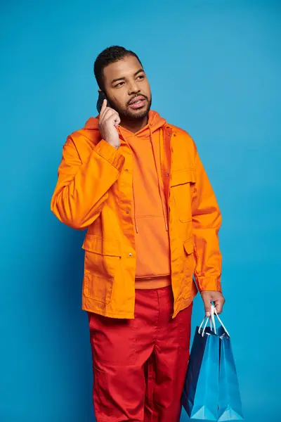Guapo afroamericano chico llamando en el teléfono inteligente y mirando a un lado con bolsas de compras - foto de stock