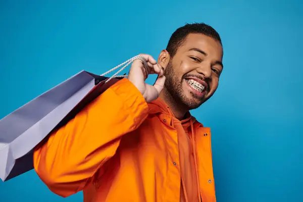 Homme afro-américain souriant en tenue orange avec sac à provisions au-dessus de l'épaule sur fond bleu — Photo de stock