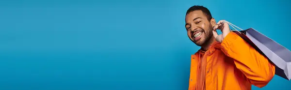 Banner of african american man in orange outfit with shopping bag over shoulder on blue background — Stock Photo