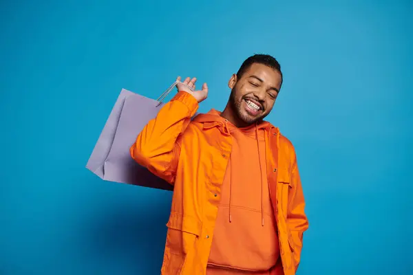 Joyful african american man in orange outfit posing with shopping bag over shoulder and closed eyes — Stock Photo