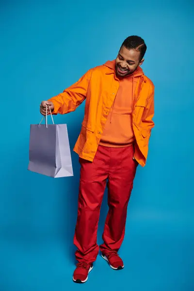Atractivo hombre afroamericano en traje naranja celebración y mirando a la bolsa de compras - foto de stock