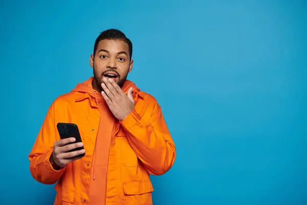 Здивований афроамериканський чоловік в помаранчевому вбранні прокручує в соціальних мережах і прикриває руку до рота — стокове фото