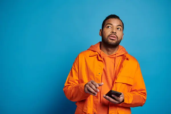 Hombre afroamericano guapo en traje naranja desplazándose en las redes sociales y mirando hacia arriba - foto de stock
