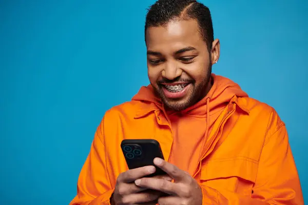 Gioioso afro americano uomo in abito arancione gioco in smartphone su sfondo blu — Foto stock
