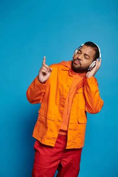 Sonriente hombre afroamericano en traje naranja y auriculares escuchando música sobre fondo azul - foto de stock
