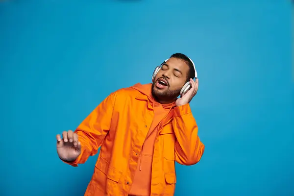 Afrikanisch-amerikanischer Mann in orangefarbenem Outfit und Kopfhörer singt mit geschlossenen Augen auf blauem Hintergrund — Stockfoto