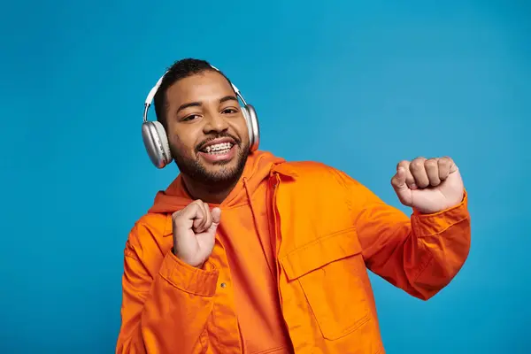 Alegre afroamericano hombre en auriculares bailando y mirando a la cámara sobre fondo azul - foto de stock