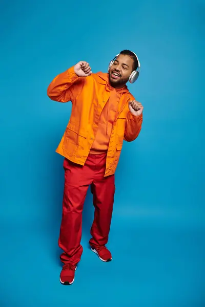 Playful african american man in orange outfit and headphones dancing on blue background — Stock Photo