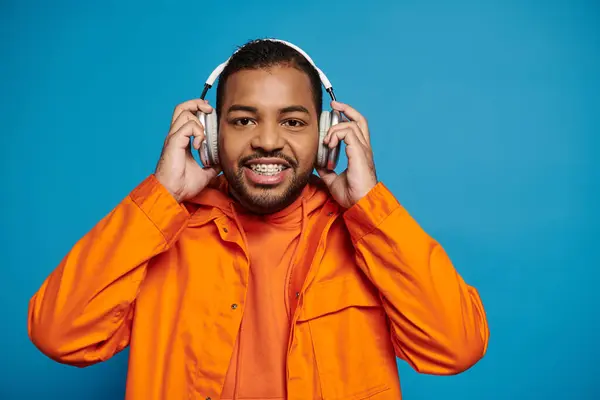 Hombre afroamericano guapo en traje naranja mirar a la cámara y sostener con las manos en los auriculares - foto de stock