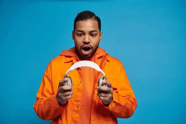 Hombre afroamericano sorprendido en traje naranja sosteniendo con las manos auriculares sobre fondo azul - foto de stock
