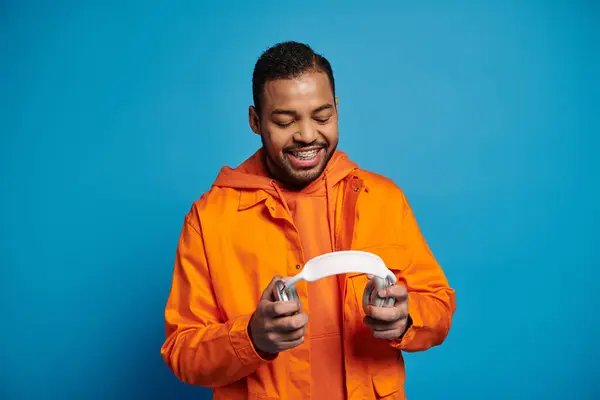 Sonriente hombre afroamericano en traje naranja sosteniendo con las manos auriculares sobre fondo azul - foto de stock