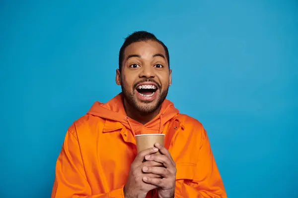 Joyeux jeune homme afro-américain en tenue orange tenant avec les mains tasse en papier sur fond bleu — Photo de stock