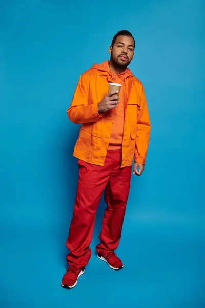 Hombre afroamericano en traje naranja de pie con taza de papel en la mano sobre fondo azul - foto de stock