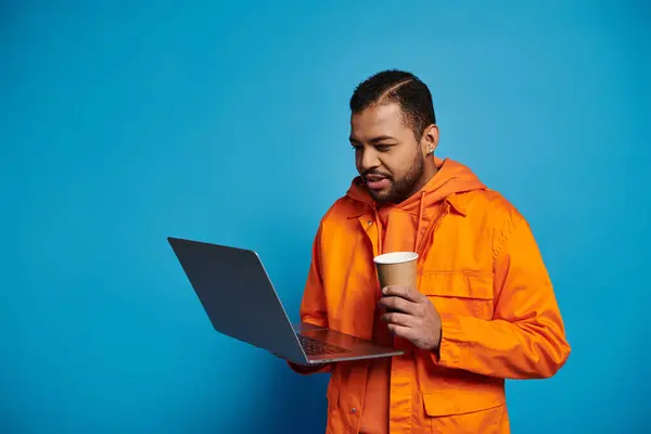 Schöner afrikanisch-amerikanischer junger Mann in orangefarbenem Outfit mit Pappbecher und Laptop auf blauem Hintergrund — Stockfoto