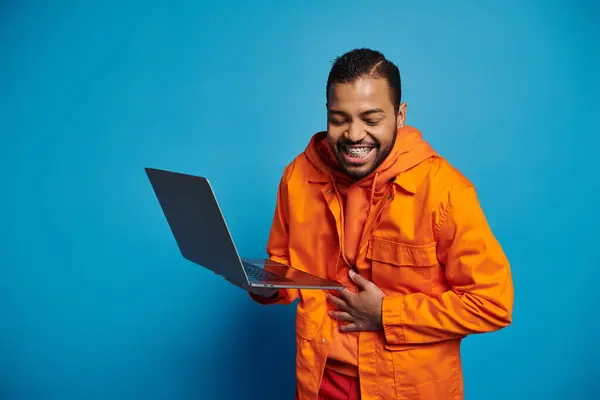 Charming african american young man in orange outfit holding laptop and laughing on blue background — Stock Photo