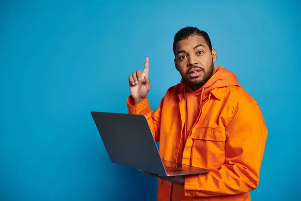 Confused african american man in orange outfit with laptop came up with idea on blue background — Stock Photo