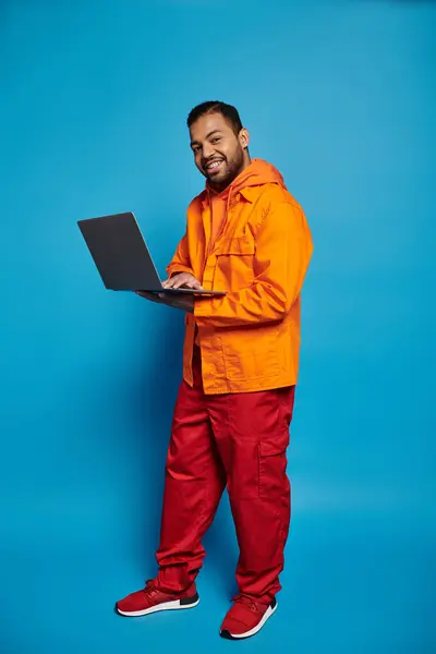 Smiling african american man in orange outfit standing sideways with laptop in hands — Photo de stock