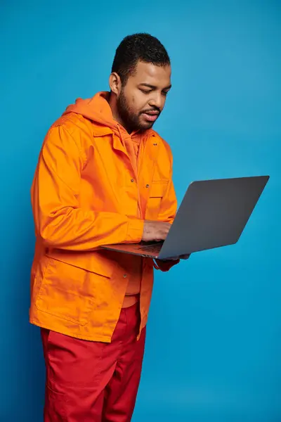 Handsome african american man in orange outfit standing and networking in laptop on blue background — Stock Photo