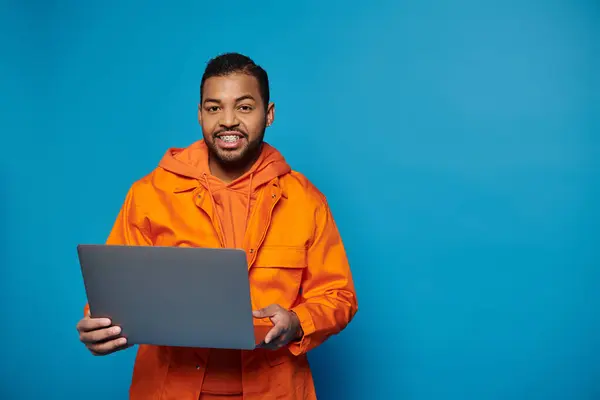 Cheerful african american man in orange outfit standing with laptop in hands and looking to camera — Stock Photo