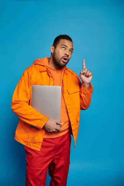 Nachdenklicher afrikanisch-amerikanischer Mann in orangefarbenem Outfit posiert mit Laptop und erhobenem Zeigefinger — Stockfoto