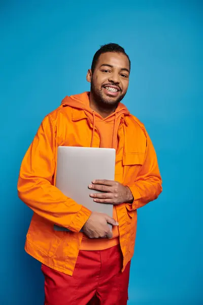 Smiling african american young guy in orange outfit posing with laptop against blue background — Photo de stock