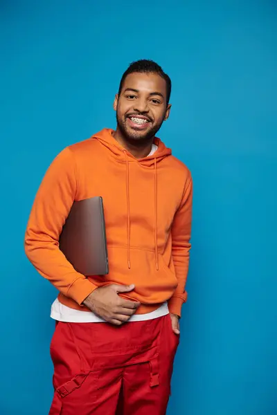 Привабливий афроамериканський хлопець у яскравому вбранні позує з ноутбуком під рукою на синьому фоні — стокове фото