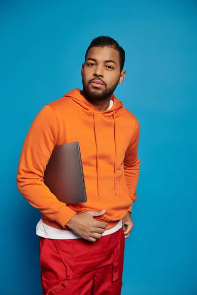 Charismatic african american man in vibrant outfit posing with laptop under arm on blue background — Stock Photo
